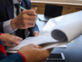 A businessman uses his pen to sign a document held by a businesswoman.